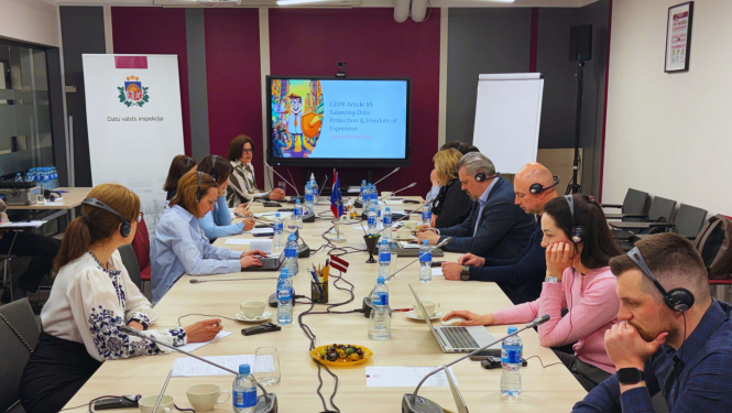 A meeting in a conference room where several participants are seated around a long table, wearing headsets and working on laptops.
