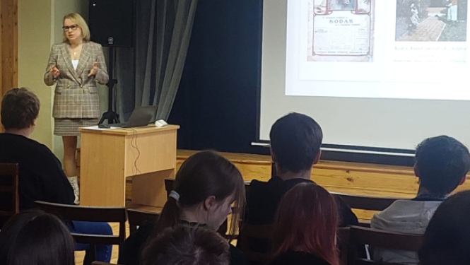 Datu valsts inspekcijas pārstāve lasa lekciju skolēniem par drošību digitālajā vidē. Viņa stāv pie galda ar datoru un prezentē materiālus uz ekrāna, kamēr skolēni uzmanīgi klausās zālē.