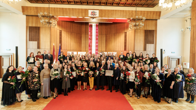 Tieslietu ministrijas zāle svinīgā apbalvošanas pasākumā, kur lielā cilvēku grupa stāv uz sarkanā paklāja, daudziem turot rokās ziedus un apbalvojumus.