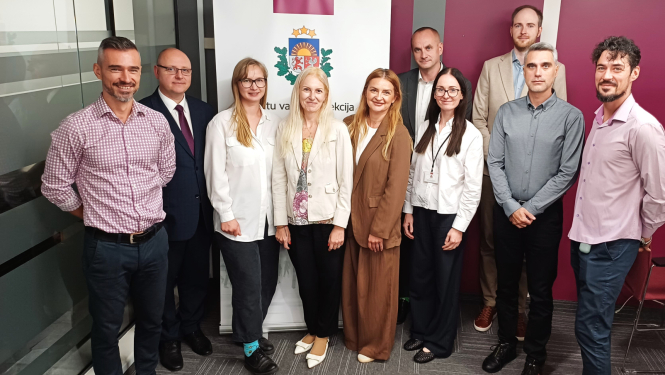 Grupas fotogrāfija ar Datu valsts inspekcijas un Rumānijas Iekšlietu ministrijas delegācijas pārstāvjiem pieredzes apmaiņas vizītes laikā, stāvot pie DVI emblēmas banera biroja telpās