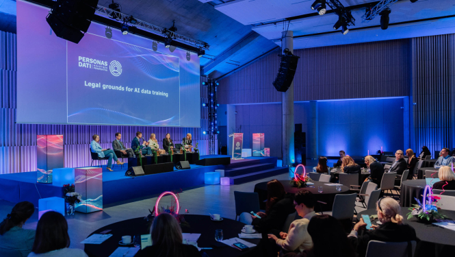 Konferences zāle ar skatu uz skatuvi, kur notiek paneļdiskusija par mākslīgā intelekta datu apstrādes juridiskajiem pamatiem. Uz skatuves sēž vairāki runātāji, bet auditorija klausās pie apaļiem galdiem. Telpa izgaismota zilganos toņos, ar lielu ekrānu fonā, kur redzams prezentācijas nosaukums.