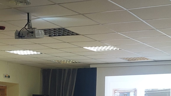 Datu valsts inspekcijas pārstāve lasa lekciju skolēniem par drošību digitālajā vidē. Viņa stāv pie galda ar datoru un prezentē materiālus uz ekrāna, kamēr skolēni uzmanīgi klausās zālē.