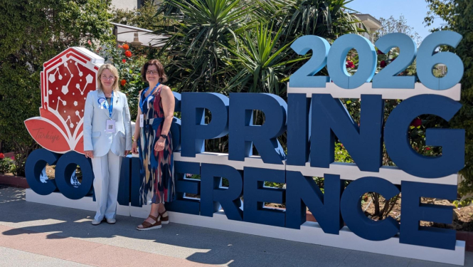 Divas sievietes stāv ārtelpā pie liela uzraksta “Spring Conference 2026”