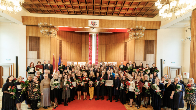 Tieslietu ministrijas zāle svinīgā apbalvošanas pasākumā, kur lielā cilvēku grupa stāv uz sarkanā paklāja, daudziem turot rokās ziedus un apbalvojumus.
