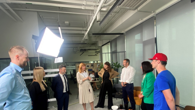 Grupas foto biroja telpās: deviņi cilvēki stāv aplī ap filmēšanas gaismu, iesaistīti sarunā vai smaidot. Telpā redzami galdi, plaukti, datori un uzkodas uz galdiņa priekšplānā.