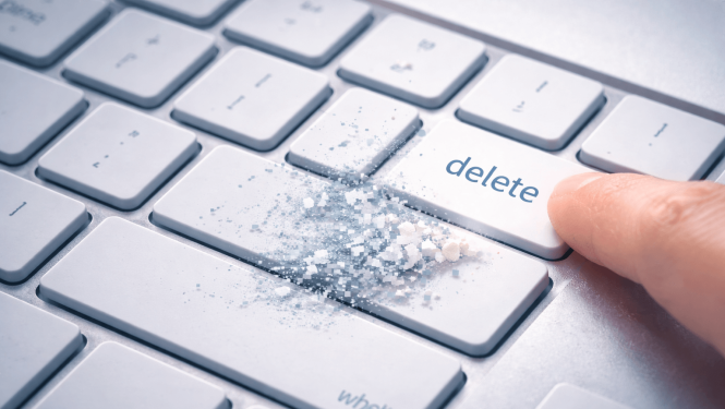 Tuvplānā redzama sudrabotas krāsas datora tastatūra, kur pirksts nospiež taustiņu ar uzrakstu “delete”.