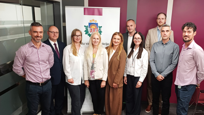 Grupas fotogrāfija ar Datu valsts inspekcijas un Rumānijas Iekšlietu ministrijas delegācijas pārstāvjiem pieredzes apmaiņas vizītes laikā, stāvot pie DVI emblēmas banera biroja telpās