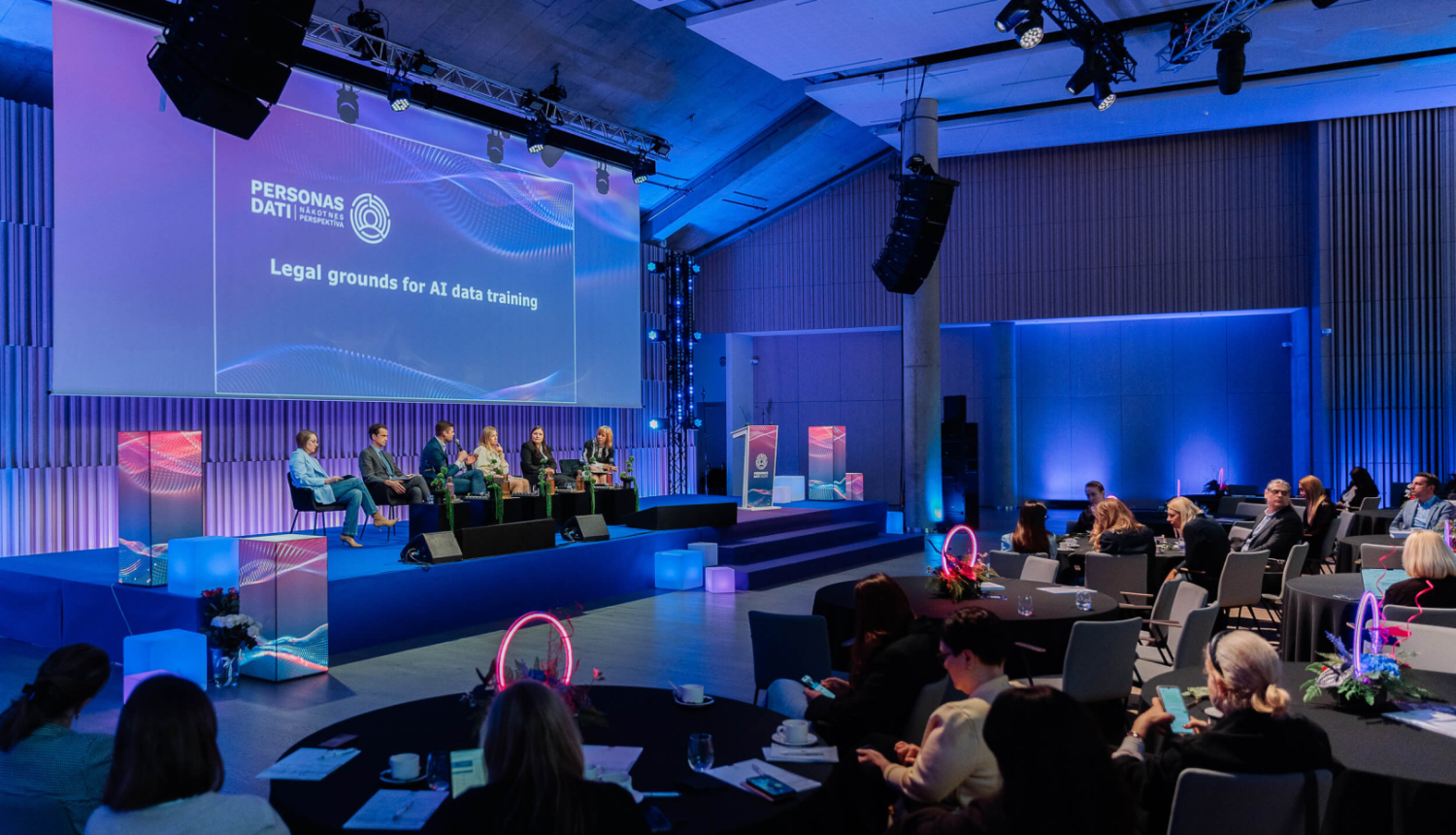Konferences zāle ar skatu uz skatuvi, kur notiek paneļdiskusija par mākslīgā intelekta datu apstrādes juridiskajiem pamatiem. Uz skatuves sēž vairāki runātāji, bet auditorija klausās pie apaļiem galdiem. Telpa izgaismota zilganos toņos, ar lielu ekrānu fonā, kur redzams prezentācijas nosaukums.