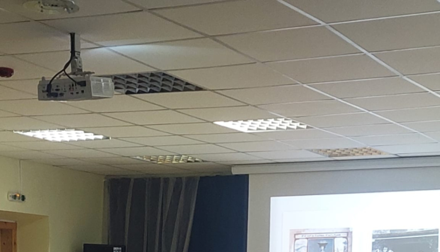 Datu valsts inspekcijas pārstāve lasa lekciju skolēniem par drošību digitālajā vidē. Viņa stāv pie galda ar datoru un prezentē materiālus uz ekrāna, kamēr skolēni uzmanīgi klausās zālē.