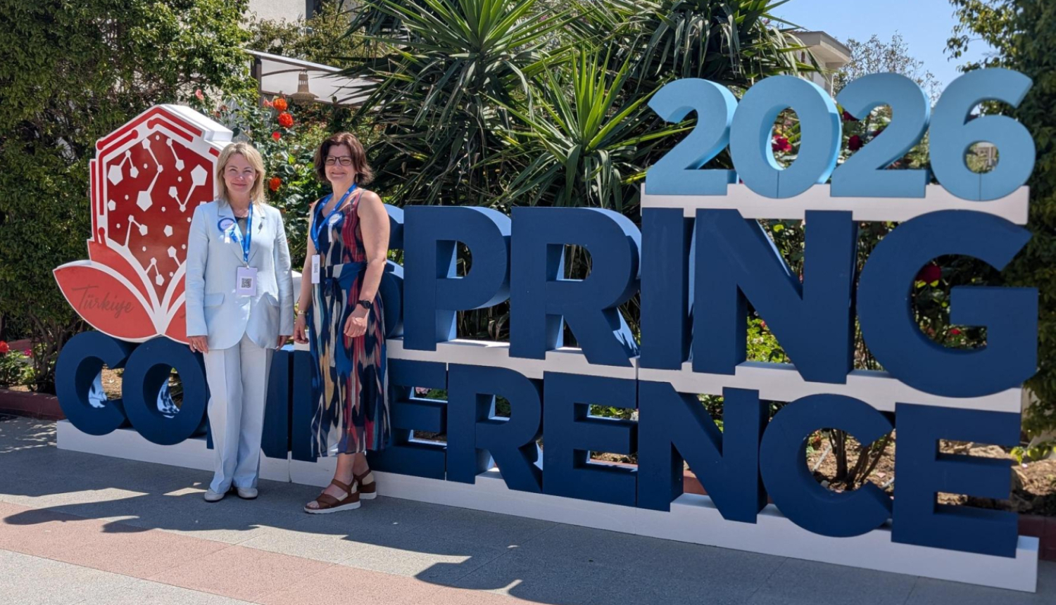 Divas sievietes stāv ārtelpā pie liela uzraksta “Spring Conference 2026”