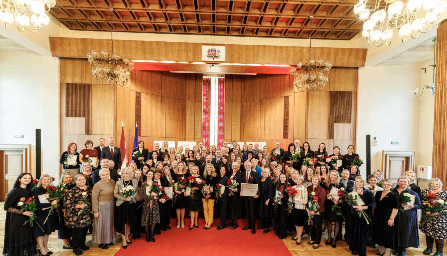 Tieslietu ministrijas zāle svinīgā apbalvošanas pasākumā, kur lielā cilvēku grupa stāv uz sarkanā paklāja, daudziem turot rokās ziedus un apbalvojumus.