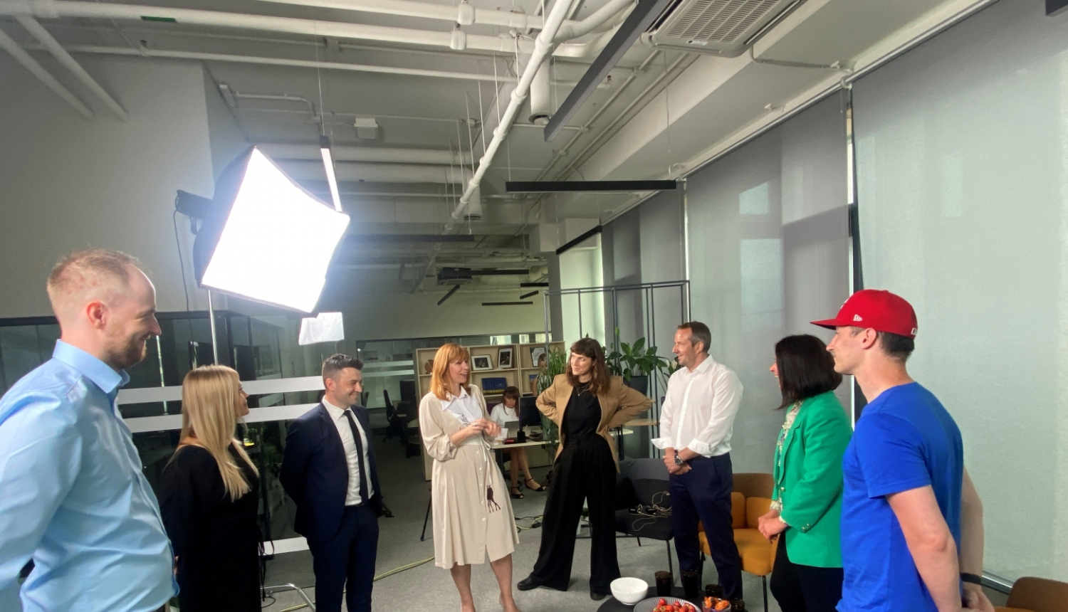 Grupas foto biroja telpās: deviņi cilvēki stāv aplī ap filmēšanas gaismu, iesaistīti sarunā vai smaidot. Telpā redzami galdi, plaukti, datori un uzkodas uz galdiņa priekšplānā.