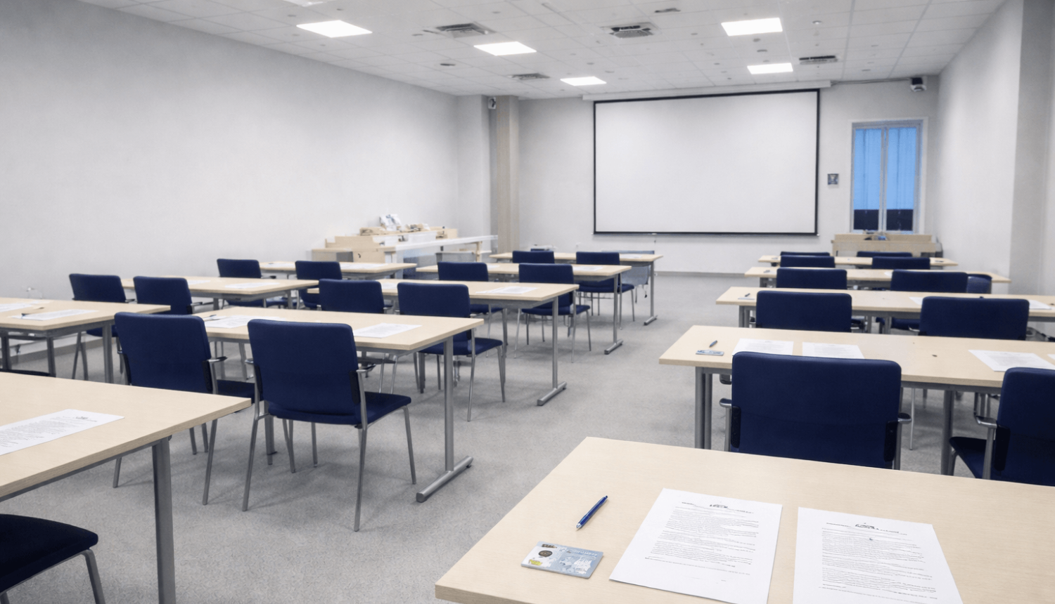 "Plaša, gaiša eksāmena telpa ar vairākām rindās izvietotām galdiem un krēsliem. Uz galdiem novietotas papīra lapas, pildspalvas un personu apliecinošs dokuments.