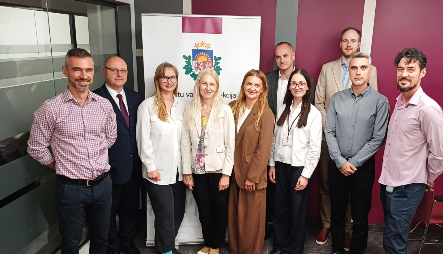 Grupas fotogrāfija ar Datu valsts inspekcijas un Rumānijas Iekšlietu ministrijas delegācijas pārstāvjiem pieredzes apmaiņas vizītes laikā, stāvot pie DVI emblēmas banera biroja telpās