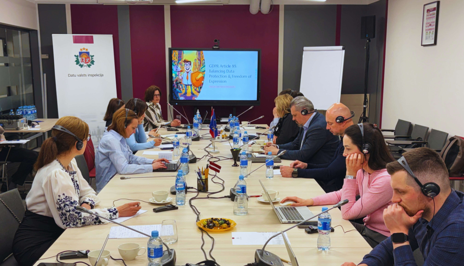 A meeting in a conference room where several participants are seated around a long table, wearing headsets and working on laptops.