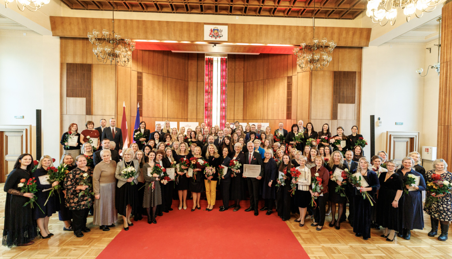 Tieslietu ministrijas zāle svinīgā apbalvošanas pasākumā, kur lielā cilvēku grupa stāv uz sarkanā paklāja, daudziem turot rokās ziedus un apbalvojumus.