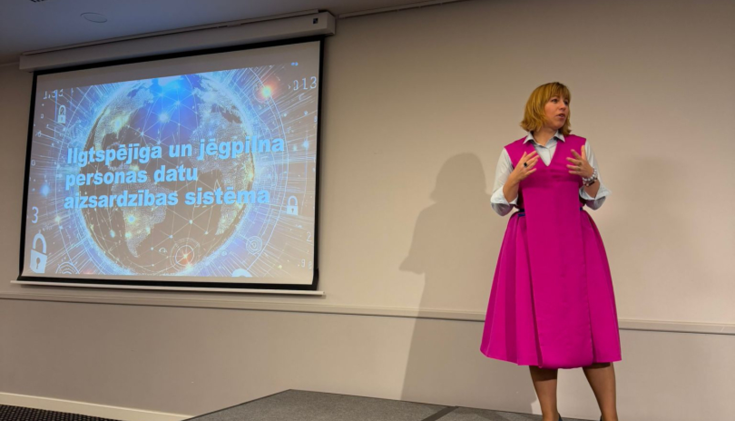 Datu valsts inspekcijas direktore uzstājas Latvijas Iekšējo auditoru institūta konferencē ar prezentāciju “Ilgtspējīga un jēgpilna personas datu aizsardzības sistēma”. Fonā redzams prezentācijas slaids ar globusa un datu tīkla vizualizāciju.